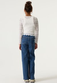 Weißes Spitzenoberteil mit langen Ärmeln und gewelltem Saum, kombiniert mit hochgeschnittenen, weiten blauen Jeans. Die Rückansicht zeigt das enganliegende Oberteil und die lockeren Jeans.
