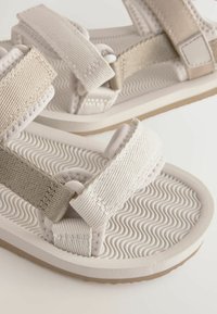 Ljust beige sandaler med vävda tygremmar, justerbara stängningar och en strukturerad gummisula med vågiga mönster.