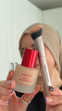 Fond de teint TIRTIR Mask Fit Red dans un flacon beige givré avec un bouchon rouge, à côté d'un pinceau à fond de teint synthétique argenté avec des poils noirs et blonds.