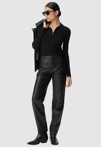 Zwart geribd longsleeve top met rits, gecombineerd met hooggetailleerde zwarte leren broek. Model draagt een zonnebril en houdt een zwart jack vast.