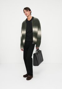 Jovem vestindo um cardigan listrado em verde e bege, camisa e calças pretas, segurando uma bolsa de couro preta, posando contra um fundo branco.