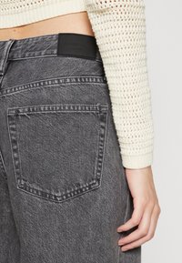 Grå denimjeans med en slät textur, som har en bakficka, läderlogotyppatch och en hög midja. Handen vilar på sidan.