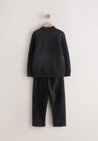 Schwarzer Pullover und passende Hose an einem Holzkleiderbügel. Glatte Textur. Einfaches Design mit gerippten Bündchen und Kragen. Keine sichtbaren Muster.