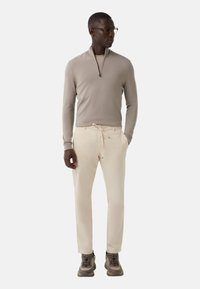 Homme portant un pull à col zippé beige, un pantalon crème à cordon, des baskets marron et des lunettes, se tenant avec une main dans sa poche devant un fond blanc.