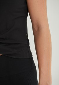 Svart ribbad linne med en sidoklyvning och en liten gyllene logotyp, parat med svarta ribbade leggings som framhäver den texturerade tyget.