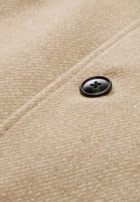 Beige wollen stof heeft een gestructureerd patroon met een donkere ronde knoop. De knoop heeft vier gaten en een glanzende afwerking.