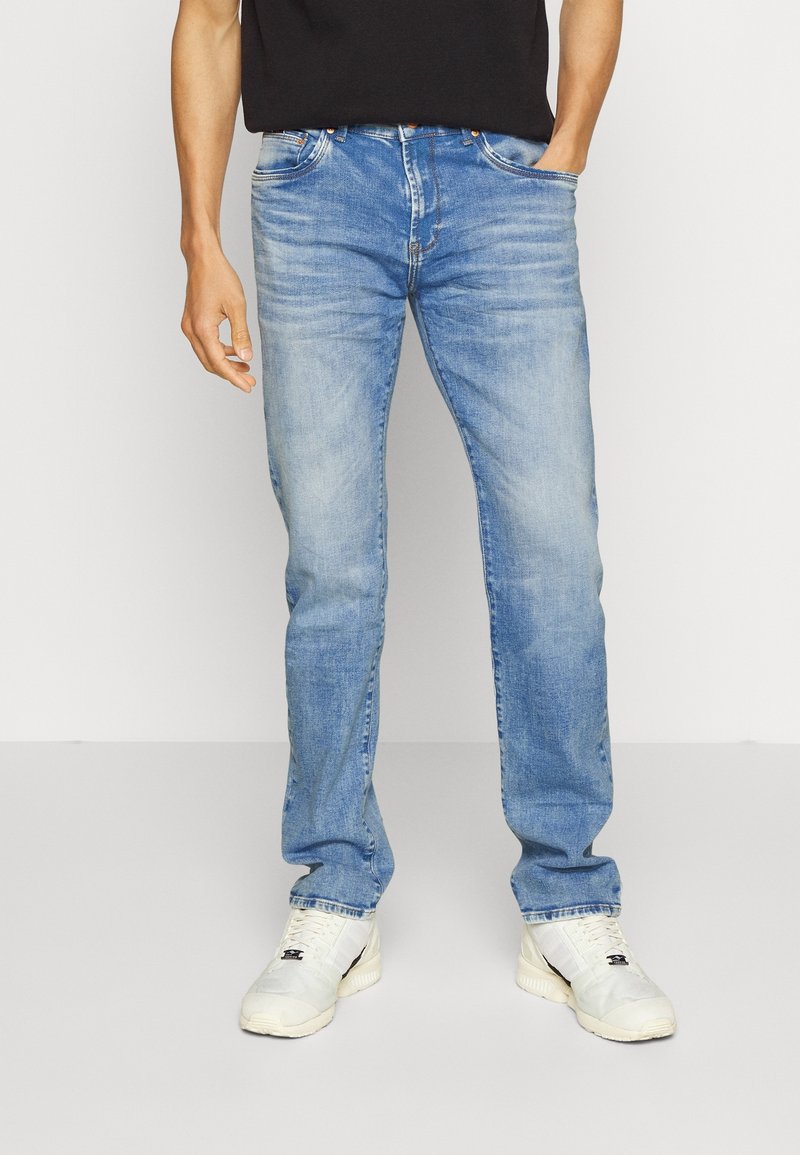 Homme portant un jean bleu clair délavé, des baskets blanches et une chemise noire, une main dans la poche, sur fond uni.