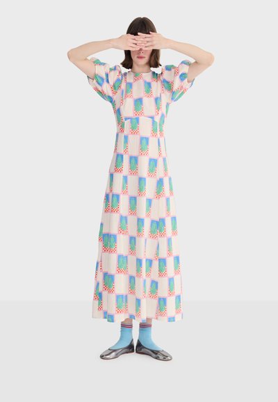 Femme portant une robe midi à motifs multicolores, chaussettes bleu clair, ballerines argentées, se couvrant les yeux avec les deux mains devant un fond blanc.