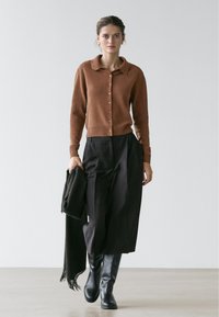 Vrouw draagt een bruin vest met knopen, zwarte broek met wijd uitlopende pijpen, zwarte laarzen, houdt een zwarte sjaal vast, loopt op een effen achtergrond.