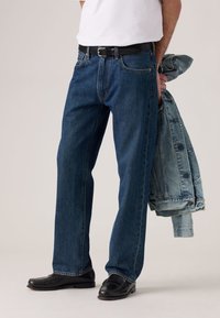 Dark blue denim jeans with a straight cut, paired with a black belt, black shoes, and light blue denim jacket draped over the arm.