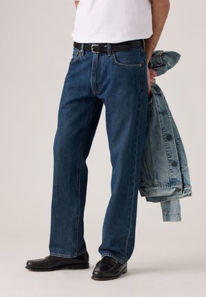 Dark blue denim jeans with a straight cut, paired with a black belt, black shoes, and light blue denim jacket draped over the arm.