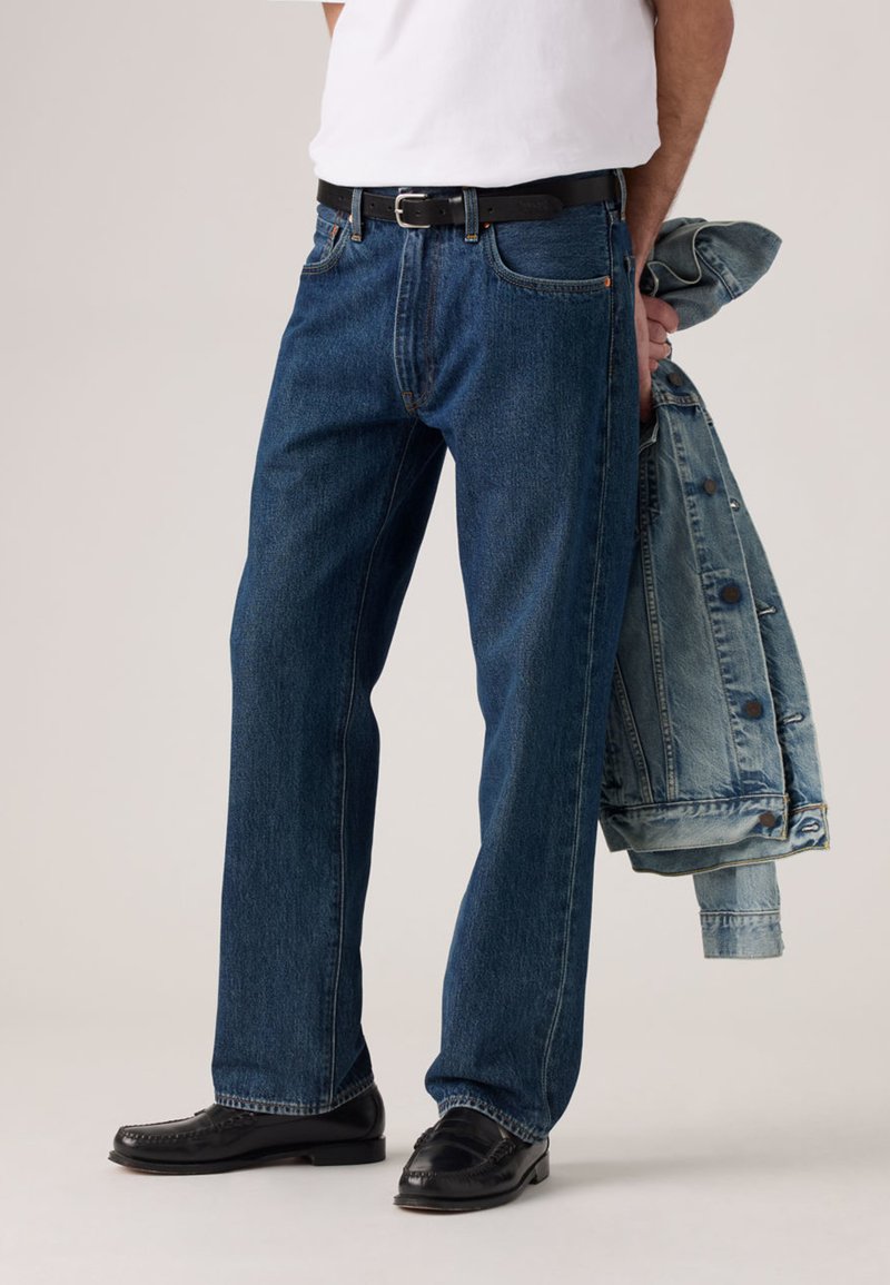 Dark blue denim jeans with a straight cut, paired with a black belt, black shoes, and light blue denim jacket draped over the arm.