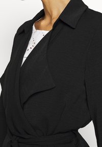 Czarny płaszcz kopertowy z kołnierzem, wykonany z teksturowanej tkaniny, z regulowanym paskiem. Przy dekolcie widoczny biały haftowany detal.