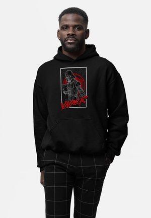 Mand iført sort hættetrøje med Darth Vader-grafik og rød "VADER" tekst, kombineret med sorte bukser med hvide gitterlinjer.