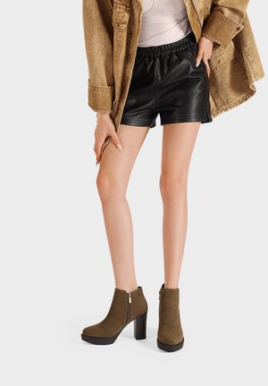 Stivali alla caviglia in suede kaki con tacco a blocco, dotati di una cerniera laterale e una suola nera liscia. Abbinati a un giubbotto marrone e pantaloni shorts in pelle nera.