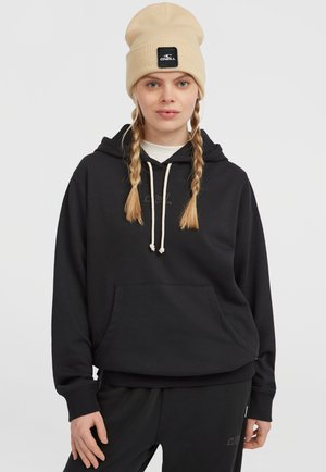 Black hoodie with a front pocket and drawstrings, made of soft material. Worn with a light-colored crew neck and a beige beanie.