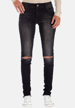 Beine in schwarzen, engen Jeans mit Rissen an den Knien, kombiniert mit weißen Sneakers und einem hellbeigen Langarmshirt, das an den Handgelenken sichtbar ist.