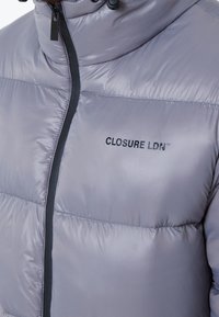 Doudoune gris clair avec fermeture éclair noire et logo "CLOSURE LDN" sur le côté gauche de la poitrine, portée par une personne à barbe visible.