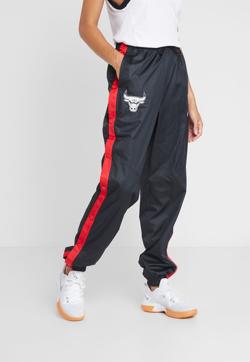 Personne portant un pantalon de sport noir avec des bandes rouges sur les côtés et le logo des Chicago Bulls, associée à des chaussures de sport blanches, debout devant un fond uni.