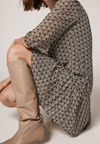 Robe à manches longues en tissu à motifs hexagonaux noirs, beiges et dorés, associée à des bottes en cuir beige montantes jusqu'au genou.