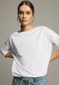 Biały, lekki, oversize'owy top z szerokim dekoltem i rękawami o długości trzech ćwierć, zestawiony z szortami dżinsowymi z wysokim stanem.