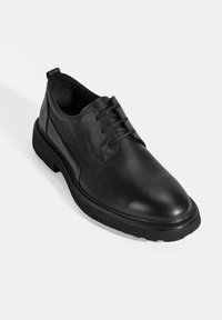 Zwarte leren schoen met een glanzende afwerking, ronde neus en traditionele vetersluiting. Voorzien van een dikke rubberen zool met textuuraccenten.