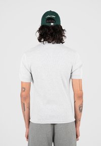 Persona con capelli lunghi che indossa un cappello verde scuro, una maglietta grigia chiara a maniche corte, pantaloni grigi e tatuaggi visibili su entrambe le braccia, di spalle.