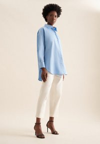 Camicia blusa azzurro chiaro con colletto a punta, abbinata a pantaloni bianchi su misura e sandali neri con tacco, su uno sfondo neutro.