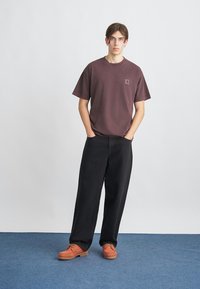 Mørkebrun bomulds t-shirt med et lille logo, parret med løse sorte denimbukser og teksturerede orange sko på en blå tæppebaggrund.