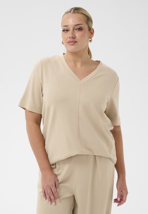 Femme aux cheveux blonds tirés en arrière, portant des boucles d'oreilles anneaux dorées, un haut beige à manches courtes et col en V, et un pantalon assorti, debout devant un fond blanc.