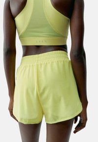 Geltonos sportinės šortai su elastine juosta, turintys lygų paviršių ir iki kelių ilgio, derinami su atitinkamu geltonu sportiniu liemenėliu.