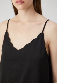 Femme portant un haut noir sans manches avec des bords festonnés et de fines bretelles, sur un fond clair uni.