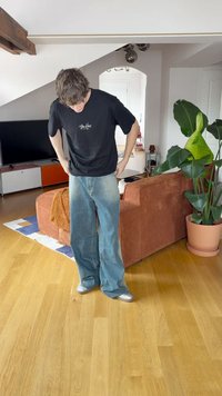 Μαύρο μπλουζάκι, ανοιχτό μπλε φαρδιά τζιν και ασημί αθλητικά παπούτσια. Στεκεται εσωτερικά με έναν καναπέ και ένα μεγάλο φυτό στο φόντο. Ξύλινο δάπεδο.