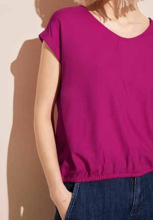 Person trägt eine kurzärmelige, magentafarbene Bluse mit elastischem Saum und dunkelblaue Jeans, steht mit einer Hand in der Tasche.