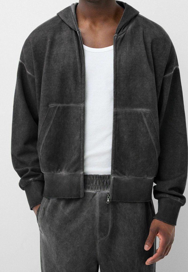 Homme portant un sweat à capuche gris foncé zippé et un pantalon de survêtement assorti avec un débardeur blanc, debout les mains dans les poches.