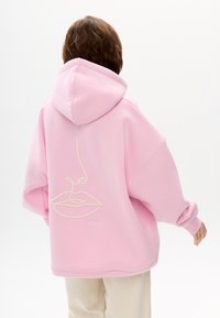 Pinkfarbener, übergroßer Hoodie mit Kordelzugkapuze. Rückseitig mit einem weißen, bestickten Design eines Gesichts und Lippen versehen. Weiche Textur und lockere Passform.