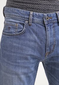 Jean en denim bleu clair à coupe droite, doté d'une poche avant, de coutures visibles et d'une fermeture à bouton métallique. Texture légèrement délavée.