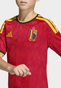 Röd barntröja för fotboll med gula detaljer och svart kantning. Har Belgiens landslagsemblem och ett mönster som liknar lågor.