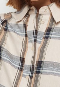 Camicia a quadri beige con chiusura a bottoni. Presenta sottili righe navy, marroni e bianche; colletto classico; tessuto morbido con una texture liscia.