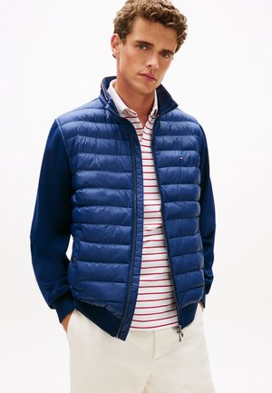 Mann trägt dunkelblaue gesteppte Jacke über weißem und rot gestreiftem Poloshirt mit beigen Hosen, Hände in den Taschen, neutraler Hintergrund.