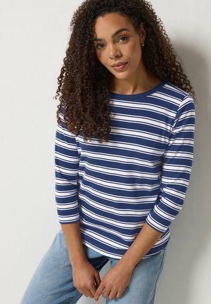 Frau mit lockigem Haar, die ein langärmliges Hemd mit marineblauen und weißen Streifen und hellblaue Jeans trägt, stehend vor einer hellen Wand.