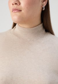 Bež polo jopica z rebrasto teksturo. Pritisnjena slika prikazuje visok ovratnik in fine pletene detajle. Preprost dizajn, minimalni šivi.