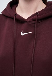 Bordo Nike kapuca z značko v belem, vrvicami na vratu in mehko, teksturirano tkanino. Poglejte bližje na obliko kapuce.