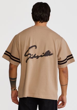 Mann mit kurzen dunklen Haaren trägt ein beiges übergroßes T-Shirt mit schwarzen Streifen an den Ärmeln und einer auffälligen schwarzen Schrift auf dem Rücken.