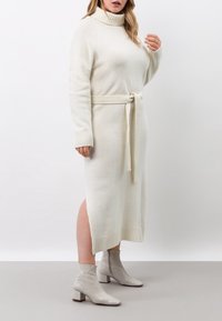 Stickad krämfärgad klänning med hög hals och knytbälte; ribbade manschetter och sidsprund. I kombination med ljusgrå texturerade ankelstövlar.