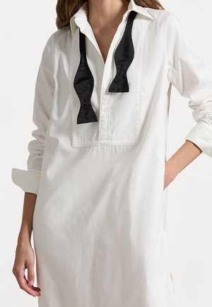 Vestido camisero blanco con detalles en negro de lazo de satén, cuello clásico, mangas largas y aberturas laterales para mayor comodidad al moverse.