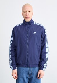Tamsiai mėlynas Adidas treniruočių švarkas su aukšta apykakle, baltais trimis juostelėmis ant rankovių ir užtrauktuku. Pagamintas iš lengvos medžiagos.