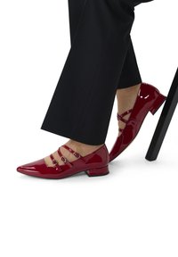 Rode lakleren schoenen met een spitse neus, drie decoratieve banden en een lage hak, gepositioneerd nabij de stoelpoten.