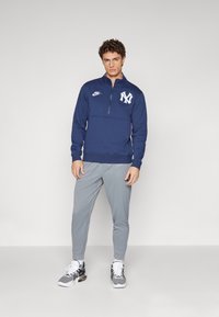 Tmavomodrý zipsový sveter s bielym logom, kombinovaný so svetlosivými zúženými nohavicami. Model má na sebe šedé a čierne tenisky.