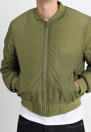 Mand iført en olivengrøn bomberjakke med lynlås, ribbet krave, ærmer og kant, hænderne i lommerne, over en hvid skjorte og mørke bukser.